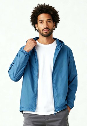 Mann richtet den Kragen einer blauen Kapuzenjacke über einem weißen T-Shirt und grauen Hosen, stehend vor einem schlichten weißen Hintergrund.