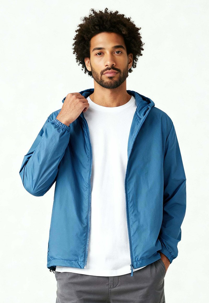 Homme ajustant le col d'une veste à capuche bleue par-dessus un t-shirt blanc et un pantalon gris, debout devant un fond blanc uni.