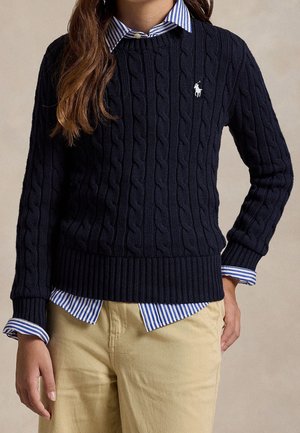 Nainen, joka pukeutuu laivastonsiniseen palmikkoneuletakkiin, sinivalkoraidallisen paidan ja beigen väristen housujen kanssa, seisten yhdellä kädellä taskussa.