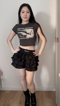 Jeune femme posant à l'intérieur, portant un crop top noir "Surfers Paradise", un short noir à volants superposé et des bottines noires.