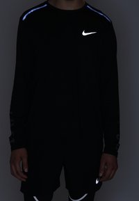 Langärmliges schwarzes Sportshirt mit reflektierendem Nike-Logo und blauen Akzenten an den Schultern, kombiniert mit schwarzen Shorts mit weißem Nike-Logo.