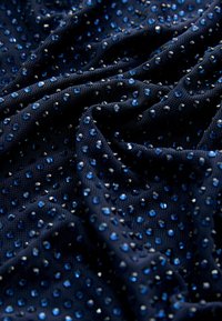 Tissu en maille marine orné de strass bleus et transparents dispersés, présentant une surface texturée et une apparence drapée et fluide.