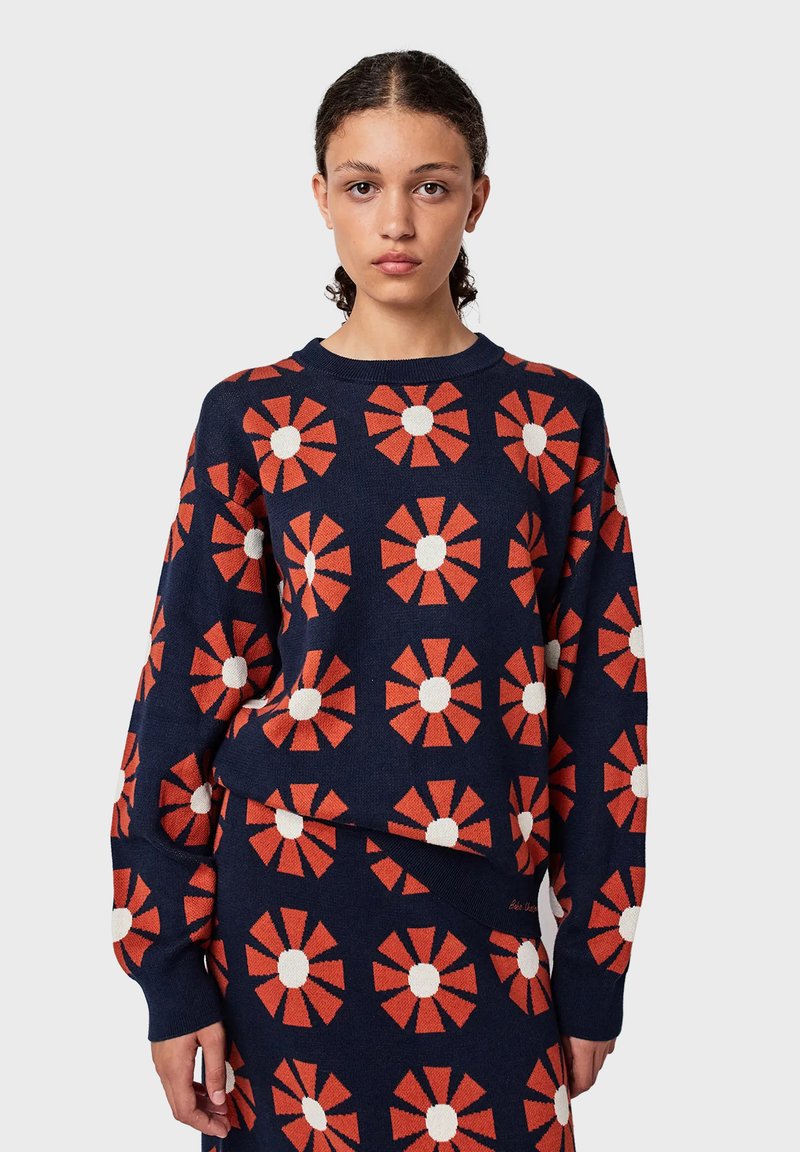 Pull en laine bleu marine avec un motif floral aux pétales orange et aux centres blancs. Col et poignets côtelés. Tissu texturé.