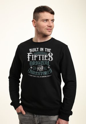 Homme aux cheveux courts et portant des boucles d'oreilles, vêtu d'un sweat-shirt noir avec le texte « Built in the Fifties Original and Unrestored », regardant sur le côté.
