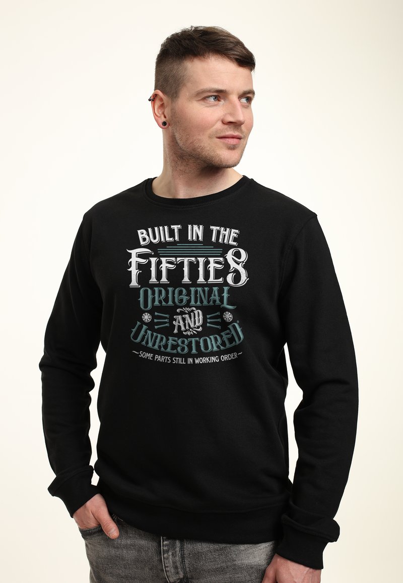 Homme aux cheveux courts et portant des boucles d'oreilles, vêtu d'un sweat-shirt noir avec le texte « Built in the Fifties Original and Unrestored », regardant sur le côté.