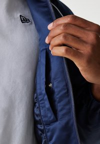 Navy blue, satin-finish jacket features a zip closure and interior pocket. A gray, soft-textured inner layer is visible, complemented by a logo.