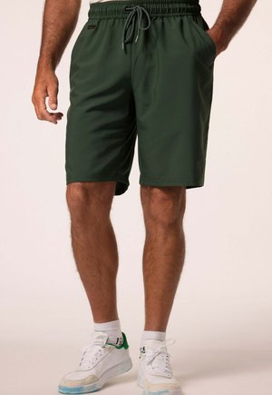 Uomo che indossa pantaloncini verde scuro con coulisse e scarpe da ginnastica bianche con accenti verdi, in piedi con le mani nelle tasche.