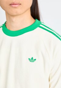 Felpa di colore crema con collo a coste verde e tre strisce verdi sulle maniche. Logo verde ricamato sul petto.