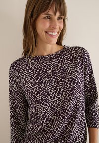 Lila topp med abstrakta vita mönster, rund halsringning och trekvartslånga ärmar. Slät tyg med en mjuk textur.