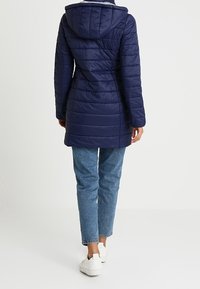 Abrigo largo acolchado azul marino con capucha, con costuras verticales y silueta entallada. Combinado con vaqueros azul claro y zapatillas blancas.