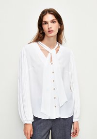 Weiße Bluse mit langen, gefältelten Ärmeln und V-Ausschnitt, ausgestattet mit einem gebundenen Kragen und goldenen Knöpfen auf der Vorderseite. Glatte Stofftextur.