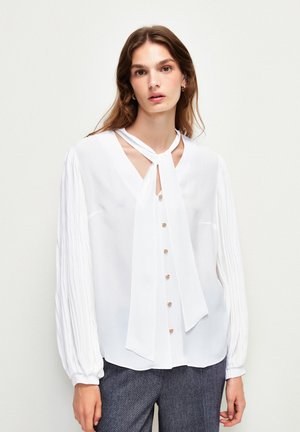 Weiße Bluse mit langen, gefältelten Ärmeln und V-Ausschnitt, ausgestattet mit einem gebundenen Kragen und goldenen Knöpfen auf der Vorderseite. Glatte Stofftextur.