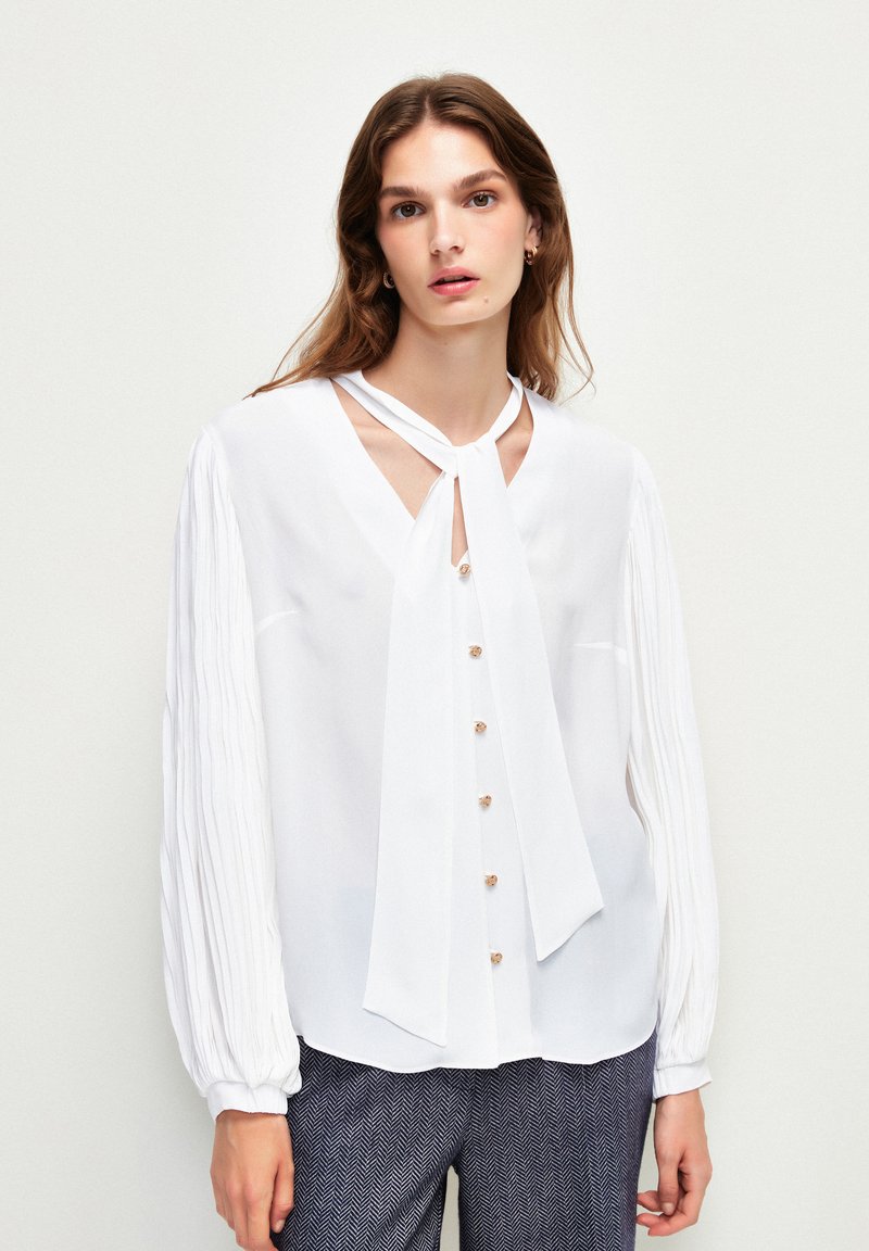 Weiße Bluse mit langen, gefältelten Ärmeln und V-Ausschnitt, ausgestattet mit einem gebundenen Kragen und goldenen Knöpfen auf der Vorderseite. Glatte Stofftextur.