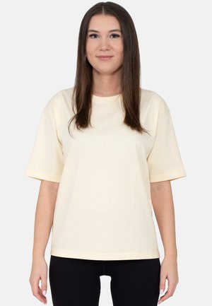 Młoda kobieta o prostych brązowych włosach nosi luźną jasnobeżową koszulkę i czarne spodnie, stoi na jednolitym białym tle.