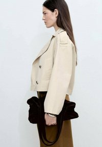 Beige kort jacka med två knappar och lapplomer; modell som håller en mörkbrun, mjuk tygväska med rynkade detaljer.