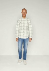 Camisa de botão de padrão xadrez claro, de mangas longas, branca com linhas pretas; combinada com jeans ajustados azuis e ténis brancos.
