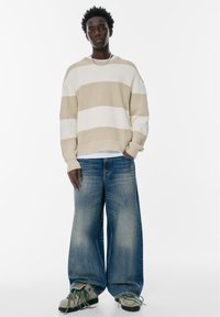 Beige en wit gestreepte gebreide trui met een geribbelde textuur; wijde, vervaagde blauwe jeans; gestructureerde grijze schoenen met groene accenten.