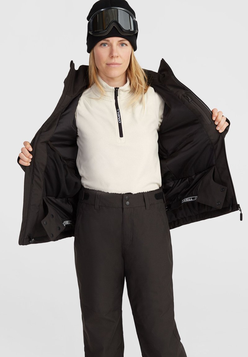 Schwarze Skijacke offen getragen, die ein cremfarbenes Fleece-Oberteil mit halblangem Reißverschluss zeigt. Schwarze Skihose mit glatter Textur und zwei Seitentaschen.