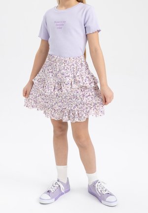 Dziewczynka w lawendowej koszulce z napisem "Shine is my favorite color", kwiecistej spódnicy z falbanami, białych skarpetkach i lawendowych tenisówkach.