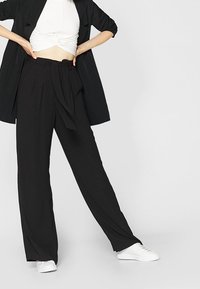 Pantalon large noir en tissu léger, avec une taille haute et une ceinture nouée, assorti à un haut blanc et des baskets blanches.