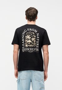 Άνδρας που φοράει μαύρο μπλουζάκι με μπεζ τροπικό σχέδιο και κείμενο "Learning From Nature Quiksilver Since 1969," συνδυασμένο με ανοιχτό μπλε τζιν.