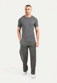 Camiseta deportiva gris de manga corta con cuello redondo, combinada con pantalones sueltos de color verde oliva. Modelo de pie con las manos en los bolsillos, vistiendo zapatillas blancas.