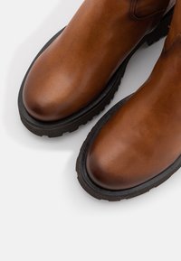 Une paire de bottes en cuir marron avec des semelles noires et robustes, vues d'en haut sur un fond blanc.