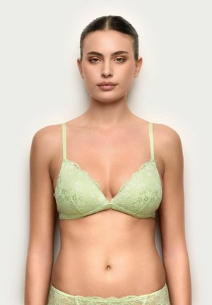 Joven mujer de frente con un sujetador de encaje verde claro y ropa interior a juego, sobre un fondo liso.
