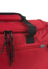 Bolsa de deporte de tela roja con una cremallera negra y dos lengüetas, que cuenta con un asa superior y un bolsillo con cremallera lateral para almacenamiento adicional.