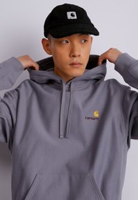 Siv hooded sweatshirt s kengursko žepom in kapuco s vrvico, na katerem je rumen in granatno rjav Carhartt logotip na prsih; nosi se z črnim klobukom.
