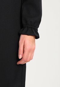 Schwarzes Kleidungsstück mit detailierter geraffter Manschette. Der Stoff wirkt glatt. Eine Hand befindet sich in der Nähe der Naht und zeigt ein natürliches Nagelfinish.