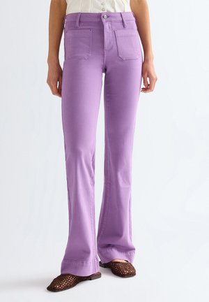 Donna che indossa jeans lavanda a vita alta con gamba dritta abbinati a scarpe piatte marroni intrecciate e una camicetta bianca senza maniche.