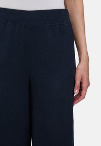 Donkerblauwe, geribde stoffen broek met een elastische tailleband. De hand rust zijwaarts en toont de gladde textuur en de losse pasvorm.