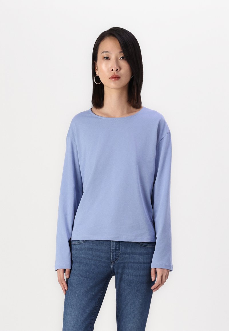 Lyseblå langermet t-skjorte i myk stoff med rund hals og avslappet passform, kombinert med mørkeblå jeans. Modellen står.