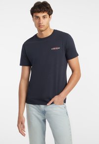 Námornícky modré bavlnené tričko s okrúhlym výstrihom, krátkymi rukávmi a malým červenobielym logom "Guess Jeans" na hrudi.