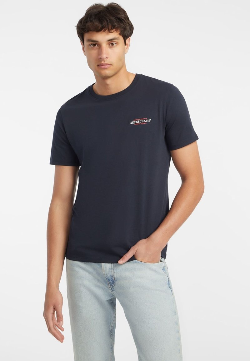 Sötétkék pamut póló kerek nyakkal, rövid ujjú, a mellen egy kis piros és fehér "Guess Jeans" logóval.