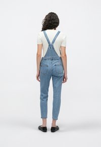 Blå denim-overalls med justerbare stropper, rett ben og to baklommer. Brukes over en hvit t-skjorte, kombinert med svarte sko.