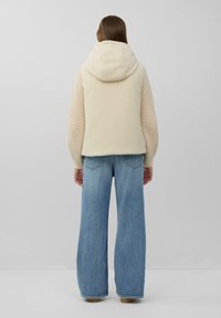 Frau, die von hinten zu sehen ist, trägt eine cremefarbene Kapuzenfleeceweste, beigen Strickärmel und weite blaue Jeans und steht vor einem schlichten grauen Hintergrund.