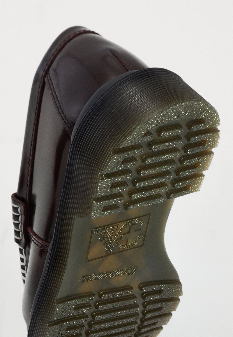 Chaussure en cuir noir brillant inclinée pour montrer la semelle en caoutchouc translucide texturée avec logo embossé et motif pour l'adhérence.