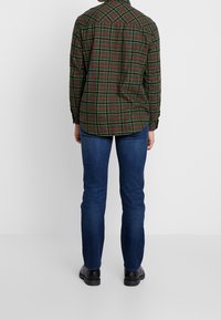 Camisa de cuadros verde con acentos rojos y amarillos, de manga larga, con dobladillo curvo, combinada con jeans azul oscuro y zapatos negros.