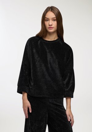 Giovane donna con lunghi capelli castani che indossa un top long-sleeve nero in velluto morbido e pantaloni abbinati, su uno sfondo semplice.