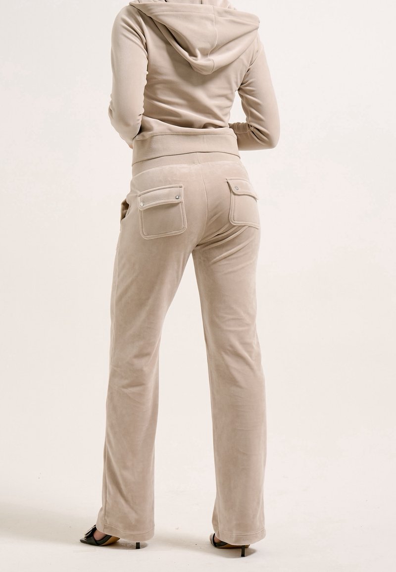 Personne portant un pantalon en velours beige avec des poches arrière à rabat et une veste à capuche assortie, debout, dos au spectateur.