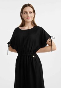 Czarna sukienka wykonana z lekkiego materiału, z okrągłym dekoltem, krótkimi rękawami z troczkami oraz elastyczną talią z złotym akcentem.