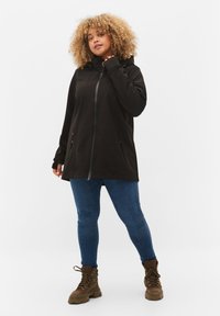 Woman with curly hair wearing a black zip-up jacket, blue jeans, and brown lace-up boots standing against a white background.