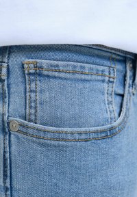 Nahaufnahme einer Tasche einer blauen Jeans mit sichtbaren Nähten und Metallniete, getragen mit einem weißen Oberteil.