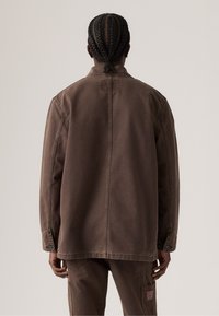 Persona con trenzas que lleva una chaqueta marrón de manga larga y pantalones a juego, de pie de espaldas contra un fondo liso.