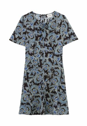 Mujer que lleva un vestido azul y negro con estampado paisley y un suéter a rayas granate atado a la cintura, sosteniendo un bolso beige, de pie sobre un fondo blanco.