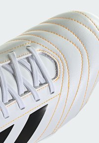 Hvide læderskolesko med orange syninger, der har en struktureret overdel, flade hvide snørebånd og et fremtrædende sort logo.