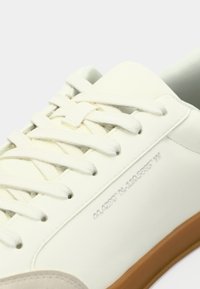 Λευκός αθλητικός παπούτσι με εκρού κορδόνια και ανοιχτό καφέ σόλα, εμφάνιση σε κοντινό πλάνο του πλαινού και πάνω μέρους με διακριτική εκτυπωμένη λεπτομέρεια κειμένου.