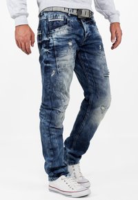 Denim jeans in various shades of blue with a distressed design, featuring rips, front pockets, and a belt, paired with white sneakers.